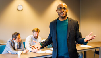 Docent in lokaal gebaart naar de camera